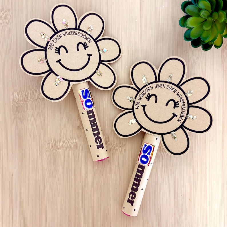 Pu&ograve; includere: Due sagome di cartone a forma di fiore color beige con contorni neri e un volto sorridente. Ogni fiore &egrave; fissato a un tubo cilindrico con l'etichetta "sommer" e una barretta di cioccolato blu e bianca. I petali presentano piccole paillettes a stella.