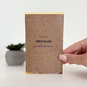 Peut inclure: Une carte rectangulaire marron avec le texte "Für den 30ER GLOW (Erhaltmaßnahme)" en noir. La carte comporte de petits points noirs et est tenue par une personne. Une petite plante succulente dans un pot gris est en arrière-plan.