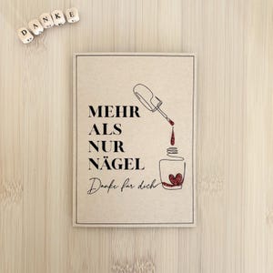 Puede incluir: Una tarjeta rectangular con el texto "MEHR ALS NUR NÄGEL Danke für dich" en escritura negra, con una ilustración de un frasco de esmalte de uñas con un diseño de corazón. La tarjeta está sobre una superficie marrón claro, con letras de madera que deletrean "DANKE".