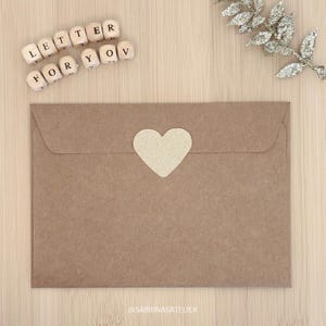 May include: A brown paper envelope with a heart-shaped sticker, with the words "LETTER FOR YOU" spelled out in wooden letter blocks. A decorative silver leaf branch is in the background. The envelope is on a light wood surface.