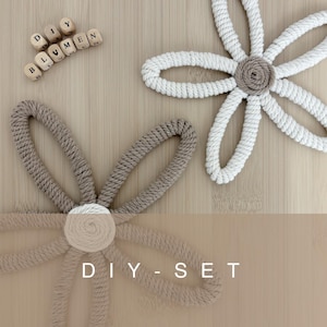 Könnte beinhalten: Zwei handgefertigte Blumen-Dekorationen aus Seil, eine weiße und eine beige, mit einem beigen Zentrum. Die Blumen befinden sich auf einer hellbraunen Holzoberfläche. Der Text "DIY - SET" steht unten. Holzbuchstaben-Perlen schreiben "BLUMEN".