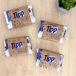 Può includere: Quattro confezioni rettangolari di snack avvolte in fascette di carta marrone con motivi di palloni da calcio. Ogni fascetta mostra il testo Tipp, un grafico di una porta e punteggi come 1:1, 1:3, 0:1 e 1:2, insieme ai nomi Lucas e Sabrinas.
