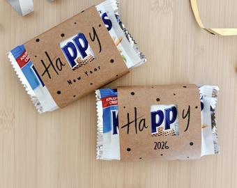 May include: Two wrapped snack bars with brown paper sleeves. The sleeves have the word "Happy" and the year "2026" or "New Year" written on them. The background is a light brown wooden surface.