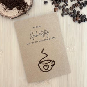 Könnte beinhalten: Eine Grußkarte aus Kraftpapier mit dem deutschen Text "Zu deinem Geburtstag" und "habe ich den Kaffeesatz gelesen". Unten befindet sich eine Kaffeetassen-Illustration mit einem Herz. Kaffeebohnen und Kaffeepulver umgeben die Karte.