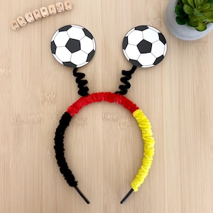 Könnte beinhalten: Ein Haarreif mit einem flauschigen Überzug in Schwarz, Rot und Gelb. Zwei schwarze Spiralfedern ragen nach oben, an deren Ende jeweils ein kreisförmiges weiß-schwarzes Fußballmotiv befestigt ist. Das Wort HAARREIF ist aus kleinen Holzbuchstaben gelegt.