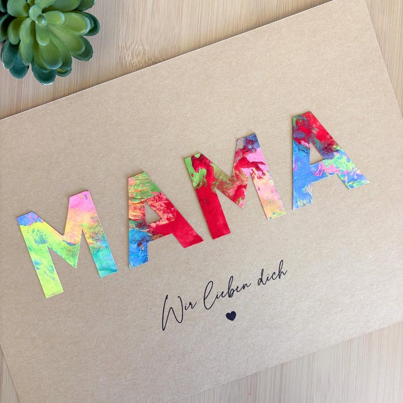 Puede incluir: Una tarjeta hecha a mano con la palabra "MAMA" en letras coloridas y abstractas. Las letras son en tonos de rosa, rojo, azul y verde. Debajo, la frase "Wir lieben dich" en cursiva, con un peque&ntilde;o coraz&oacute;n.
