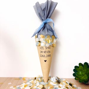 Peut inclure: Un sac cadeau conique en papier brun clair avec le texte VIEL SPAB IM NEUEN SCHULJAHR et un petit cœur noir imprimé sur le devant. Le haut est resserré par un ruban en tissu bleu et du papier crépon gris, décoré de confettis dorés.