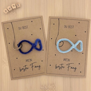 May include: Two greeting cards with a fish-shaped design made of blue and light blue pipe cleaners. The cards read "DU BIST MEIN bester Fang" and are decorated with small black hearts. Wooden letter blocks spell "LOVE".