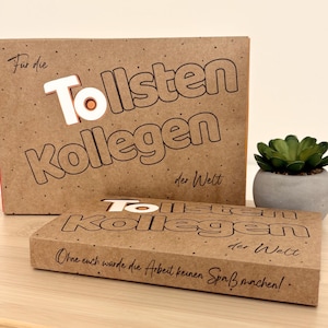 Op de afbeelding: Twee bruine kartonnen geschenkdozen met de Duitse tekst "Tollsten Kollegen der Welt". Het woord "Tollsten" is wit met een oranje cirkel in het midden. De kleinere doos heeft de tekst "Ohne euch würde die Arbeit keinen Spaß machen!"