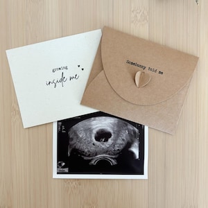 Op de afbeelding: Een zwangerschapsaankondigingsset met een echo, een crèmekleurige kaart met de tekst "growing inside me" en een bruine kaart met de tekst "Somebunny told me" en een hartvormig ornament. De items zijn gerangschikt op een licht houten oppervlak.