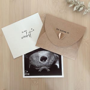 以下が含まれることがあります： 超音波画像、テキスト「in mir wächst」が書かれたクリーム色のカード、テキスト「Ein kleines Ei」と金のハートが付いた茶色の封筒が特徴の妊娠発表セット。