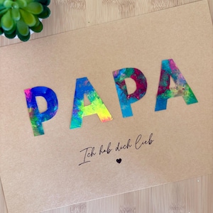 May include: A kraft paper card with the word "PAPA" in large, colorful, abstract letters. Below the word is the phrase "Ich hab dich lieb" in cursive, with a small heart symbol.