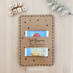 Op de afbeelding: Een bruine beterschapskaart met een gestikte rand en zwarte hartjes. De kaart bevat twee theezakjes en de tekst "Gute Besserung" en houten blokjes met "Get Well".