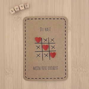 Könnte beinhalten: Eine rechteckige Karte in Braun mit einem Tic-Tac-Toe-Spiel. Rote Herzen ersetzen die Kreuze und Nullen. Der Text lautet "DU HAST MEIN HERZ EROBERT". Holzblöcke mit dem Wort "LOVE".