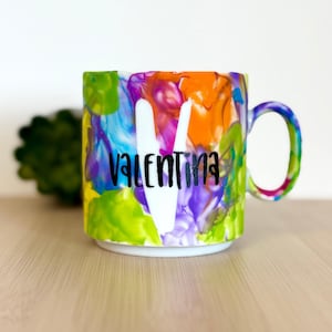 Könnte beinhalten: Ein Keramikbecher mit einem lebendigen, mehrfarbigen Marmormuster in Orange-, Lila-, Grün- und Blautönen. Ein großer weißer Buchstabe V ist mittig auf der Vorderseite platziert, überlagert von dem schwarzen kursiven Schriftzug Valentina.