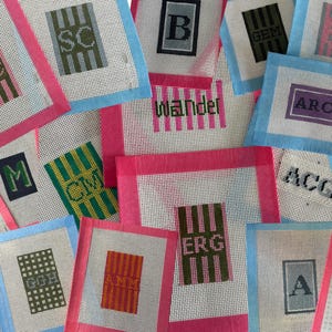 May include: A collection of needlepoint canvases featuring various monograms and letters including DC, SC, B, GEM, S, ARC, M, CM, wander, ACG, G, KR, ERG, GGH, AMM, A, and L. The canvases are bordered with pink or blue tape and display diverse striped patterns.