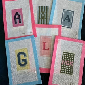 May include: Six rectangular needlepoint canvases featuring various letters and patterns. The canvases have pink or blue fabric borders. The designs include the letters A, M, A, G, L, and the letters NGK on a green grid background. Each piece is stitched.