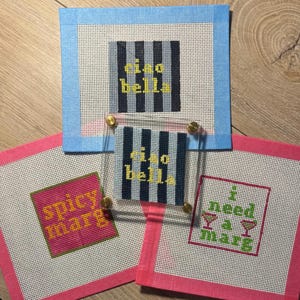 May include: Three square needlepoint canvases with taped edges. One features blue and grey stripes with the yellow text "ciao bella". Another has a pink border and the yellow text "spicy marg". The third has a pink border and the green text "i need a marg" with two cocktail glass icons. A clear acrylic frame sits over the striped canvas.