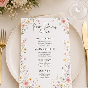 Könnte beinhalten: Eine rechteckige weiße Papiermenükarte für eine Babyparty mit einem Blumenrand aus rosa, gelben und violetten Wildblumen. Der Text lautet Baby Shower MENU, APPETIZERS, MAIN COURSE, DESSERT und DRINKS mit einer Liste von Speisen und Getränken.