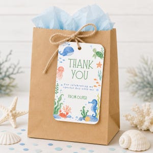 May include: A brown paper gift bag tied with twine, holding blue tissue paper. A decorative tag with a watercolor ocean theme says "Thank You" and "From Oliver". The tag features a whale, turtle, seahorse, and other sea creatures.