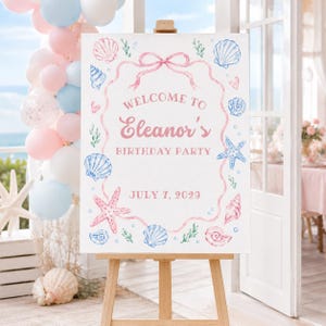 May include: A rectangular white birthday party sign on a wooden easel featuring a pink ribbon border with watercolor illustrations of blue and pink seashells, starfish, and seaweed. The text reads: WELCOME TO Eleanor's BIRTHDAY PARTY JULY 7, 2029.