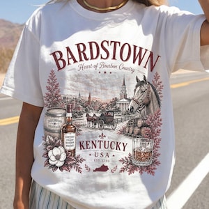 Może przedstawiać: Biały t-shirt z krótkim rękawem z nadrukiem graficznym zawierającym tekst BARDSTOWN Heart of Bourbon Country KENTUCKY USA EST. 1780. Wzór przedstawia konia, powóz konny, butelkę whisky, beczkę oraz szklankę z trunkiem.