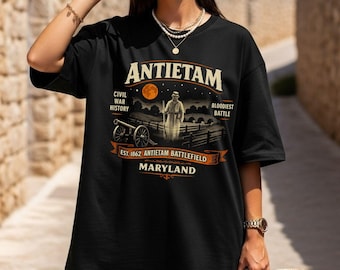 May include: Black t-shirt featuring a graphic design with the word "Antietam" at the top. The design includes an illustration of a Civil War scene with a cannon, a figure, and the text "Civil War History" and "Bloodiest Battle".