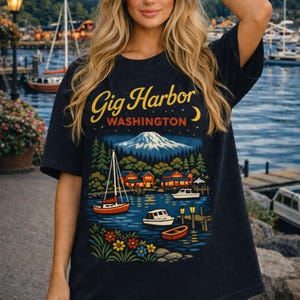 Może przedstawiać: Granatowa koszulka z malowniczą ilustracją Gig Harbor w stanie Waszyngton. Projekt zawiera górę, łodzie i napis "Gig Harbor WASHINGTON" w kolorze żółtym i białym. Koszulka ma luźny krój.