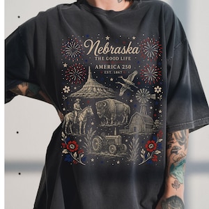 Op de afbeelding: Een antracietgrijs t-shirt met een grafische print met de tekst Nebraska The Good Life America 250 Est. 1867. Het ontwerp bevat illustraties van een cowboy te paard, een bizon, een tractor, een schuur, een windmolen, vogels en vuurwerk.
