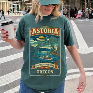 Può includere: T-shirt verde acqua con grafica del ponte Astoria-Megler, una barca e un faro. Il testo "ASTORIA" è in alto, con "PACIFIC NORTHWEST EST. 1811 OREGON" sotto. La maglietta ha una vestibilità ampia.