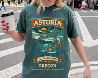 May include: Teal t-shirt featuring a graphic of the Astoria-Megler Bridge, a boat, and a lighthouse. The text "ASTORIA" is at the top, with "PACIFIC NORTHWEST EST. 1811 OREGON" below. The shirt is a relaxed fit.