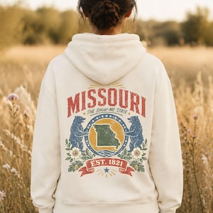 May include: A cream-colored hooded sweatshirt featuring a graphic on the back with the text MISSOURI, THE SHOW-ME STATE, and EST. 1821. The design includes a central map of Missouri, two blue bears, floral elements, and a red banner.