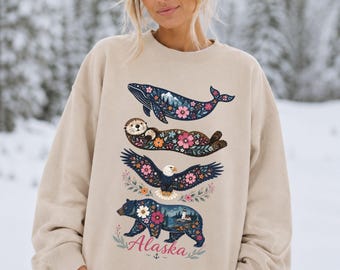 May include: A beige crewneck sweatshirt featuring a vertical graphic print of a whale, an otter, an eagle, and a bear, all decorated with floral patterns and nature scenes. The word Alaska is printed in red script at the bottom of the design.