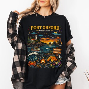 May include: Black t-shirt with a colorful graphic of Port Orford, Oregon. The design includes a lighthouse, boat, ocean waves, campfire, tent, and the text "PORT ORFORD OREGON".