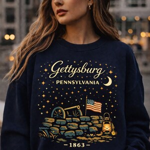 May include: Navy blue sweatshirt featuring a graphic of Gettysburg, Pennsylvania, with a cannon, American flag, and lantern. The text "Gettysburg Pennsylvania" is in gold script, with the year "-1863-" below the graphic.