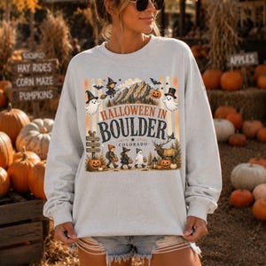 Può includere: Una felpa girocollo grigio chiaro con una stampa grafica che riporta il testo HALLOWEEN IN BOULDER COLORADO. Il design include illustrazioni di fantasmi, bambini in costume, una catena montuosa, un alce e zucche in un contesto autunnale.