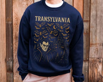 Puede incluir: Sudadera azul marino con la palabra "TRANSYLVANIA" en letras doradas. El diseño presenta murciélagos, una luna creciente, un castillo y una ilustración de vampiro. Prenda cómoda e informal.