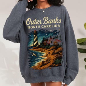 May include: A dark gray sweatshirt featuring a coastal scene with a lighthouse, beach, and the text "Outer Banks North Carolina." The graphic includes a lighthouse, ocean, and houses.
