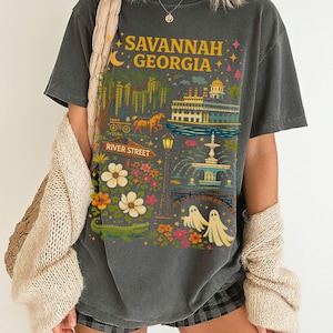 May include: Dark gray t-shirt featuring a colorful graphic of Savannah, Georgia. The design includes the text "Savannah Georgia", a horse-drawn carriage, a river scene, a fountain, flowers, and ghosts.