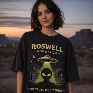May include: Black t-shirt with a graphic featuring a UFO, alien, and the text "ROSWELL NEW MEXICO - THE TRUTH IS OUT THERE." The design includes a green beam of light shining down on the alien silhouette, with cacti and a crescent moon.