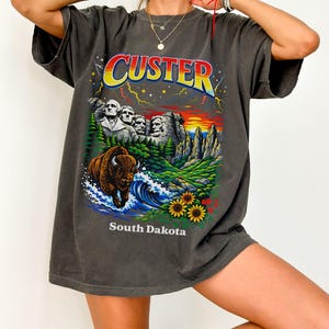 May include: Dark gray t-shirt featuring a colorful graphic of Mount Rushmore, a bison, and the text "Custer South Dakota." The design includes a sunset, waves, and sunflowers. The shirt has short sleeves and a relaxed fit.