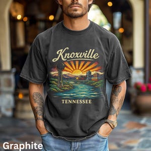 Puede incluir: Camiseta de color grafito con un diseño de Knoxville, Tennessee. El gráfico incluye una puesta de sol, un río, un puente y la Sunsphere de Knoxville. El texto "Knoxville" está arqueado sobre la imagen, con "Tennessee" debajo.