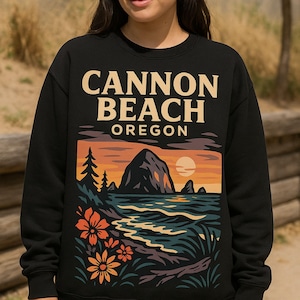 Peut inclure: Sweat-shirt noir avec un motif de Cannon Beach, Oregon. Le dessin représente un coucher de soleil sur la mer avec une grande formation rocheuse, des arbres et des fleurs. Le texte "CANNON BEACH OREGON" est imprimé en haut du motif.
