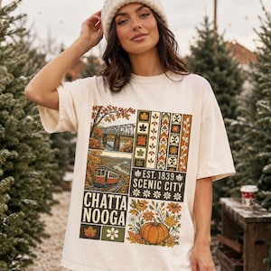 Puede incluir: Una camiseta de manga corta color hueso con un estampado gráfico que muestra un puente, un tranvía, una calabaza y hojas de otoño. El texto dice CHATTANOOGA y EST. 1839 SCENIC CITY. El diseño incluye patrones geométricos y motivos florales en tonos tierra.