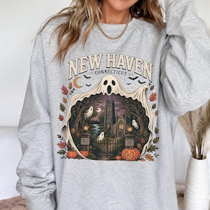 Op de afbeelding: Een lichtgrijze sweater met ronde hals en een grafische print van een geest over een begraafplaatsscène met de tekst New Haven Connecticut. Het ontwerp bevat pompoenen, herfstbladeren, vleermuizen, een halve maan en een gotisch gebouw.