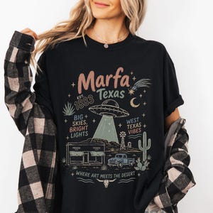 Może przedstawiać: Czarny t-shirt z nadrukiem graficznym z tekstem Marfa Texas Est. 1883, Big Skies, Bright Lights, West Texas Vibes oraz Where Art Meets The Desert. Wzór zawiera latający spodek, budynek, ciężarówkę i kaktusa.