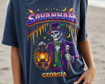 May include: Dark blue t-shirt featuring a colorful graphic of a skeleton in a top hat holding a lantern and cane. The word "SAVANNAH" is arched across the top, with "GEORGIA" below. The design has a vintage, slightly distressed look.