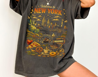 May include: Dark gray t-shirt with a colorful graphic of New York City landmarks, including the Statue of Liberty, skyline, and Broadway. The text "Welcome to NEW YORK" is arched across the top. Other elements include pizza, a pretzel, and a coffee cup.