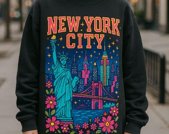 May include: Black sweatshirt featuring a colorful graphic of the New York City skyline, the Statue of Liberty, and the text "NEW YORK CITY". The design includes pink, blue, and yellow accents, with floral elements at the bottom.