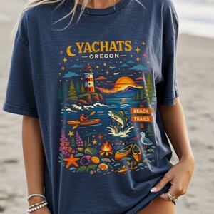 May include: Navy blue t-shirt featuring a colorful graphic of Yachats, Oregon. The design includes a lighthouse, ocean scene with a kayaker and jumping fish, and the text "Yachats Oregon". The shirt has a relaxed fit.